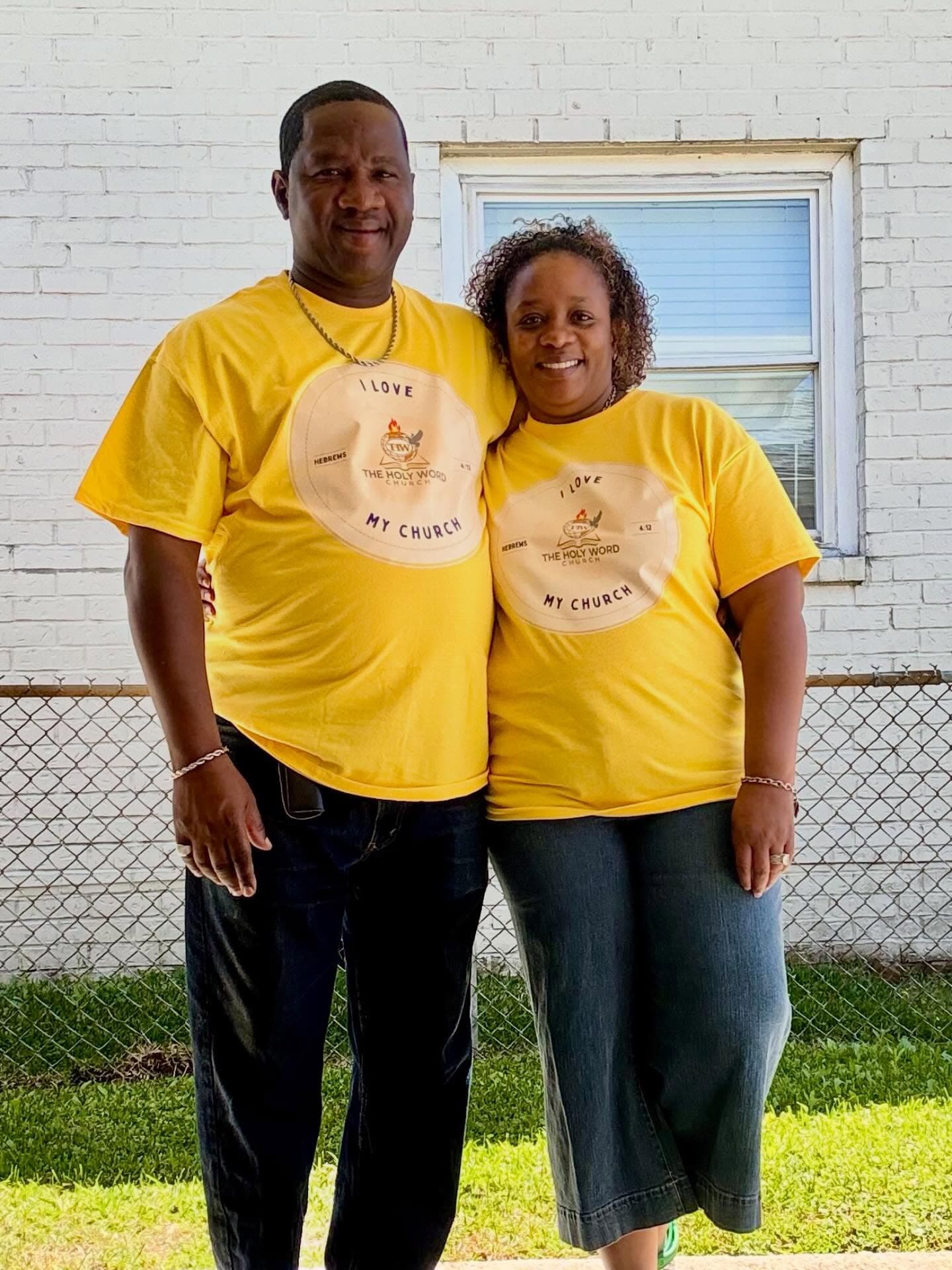 Pastor Charlie Grear Jr. and Lady Terri Grear in yellow “I love my church” shirts beside the white brick wall of the church