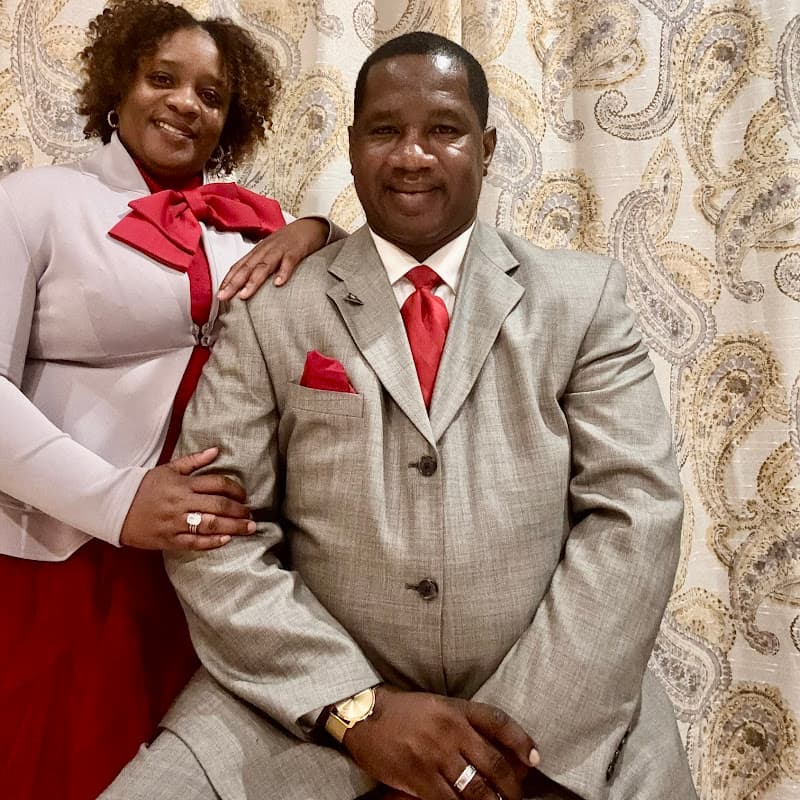 Pastor Charlie Grear Jr. in a grey suit and red tie, seated, with Lady Terri Grear standing beside him